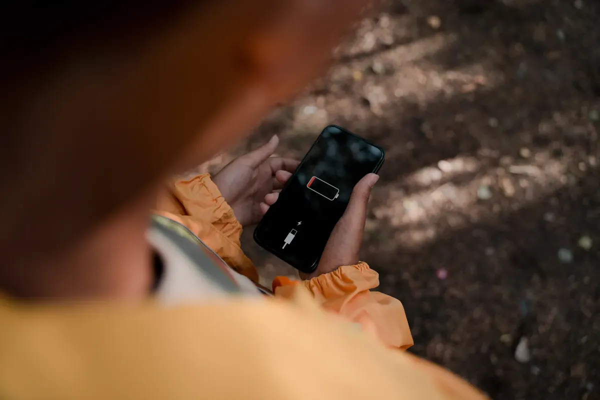 Teenage boy holds a smartphone displaying low battery outdoors in a forest.