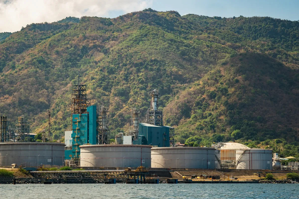 Industrial plant with large tanks and chimneys against a lush mountain backdrop.