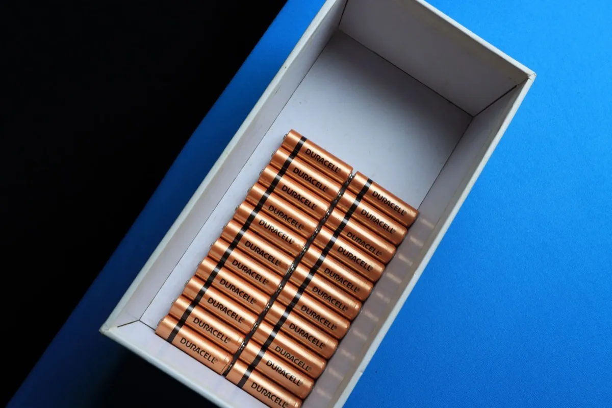Una scatola bianca aperta su un tavolo blu contiene file di pile arancioni Duracell con fascia nera, lasciando vuota la parte superiore.