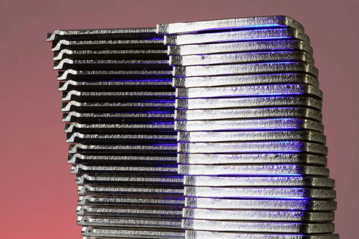 Een hoge stapel dunne zilveren metalen stroken met kleine inkepingen glanst paars en blauw tegen een zachte roze achtergrond.