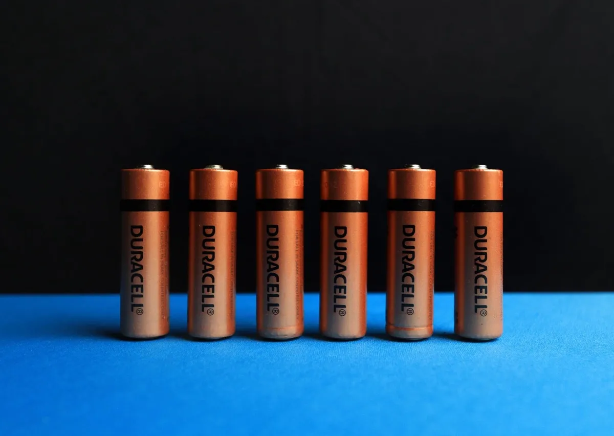 Seis pilhas cilíndricas cor de cobre da Duracell ficam alinhadas sobre uma superfície azul, com uma faixa preta e fundo escuro atrás delas.